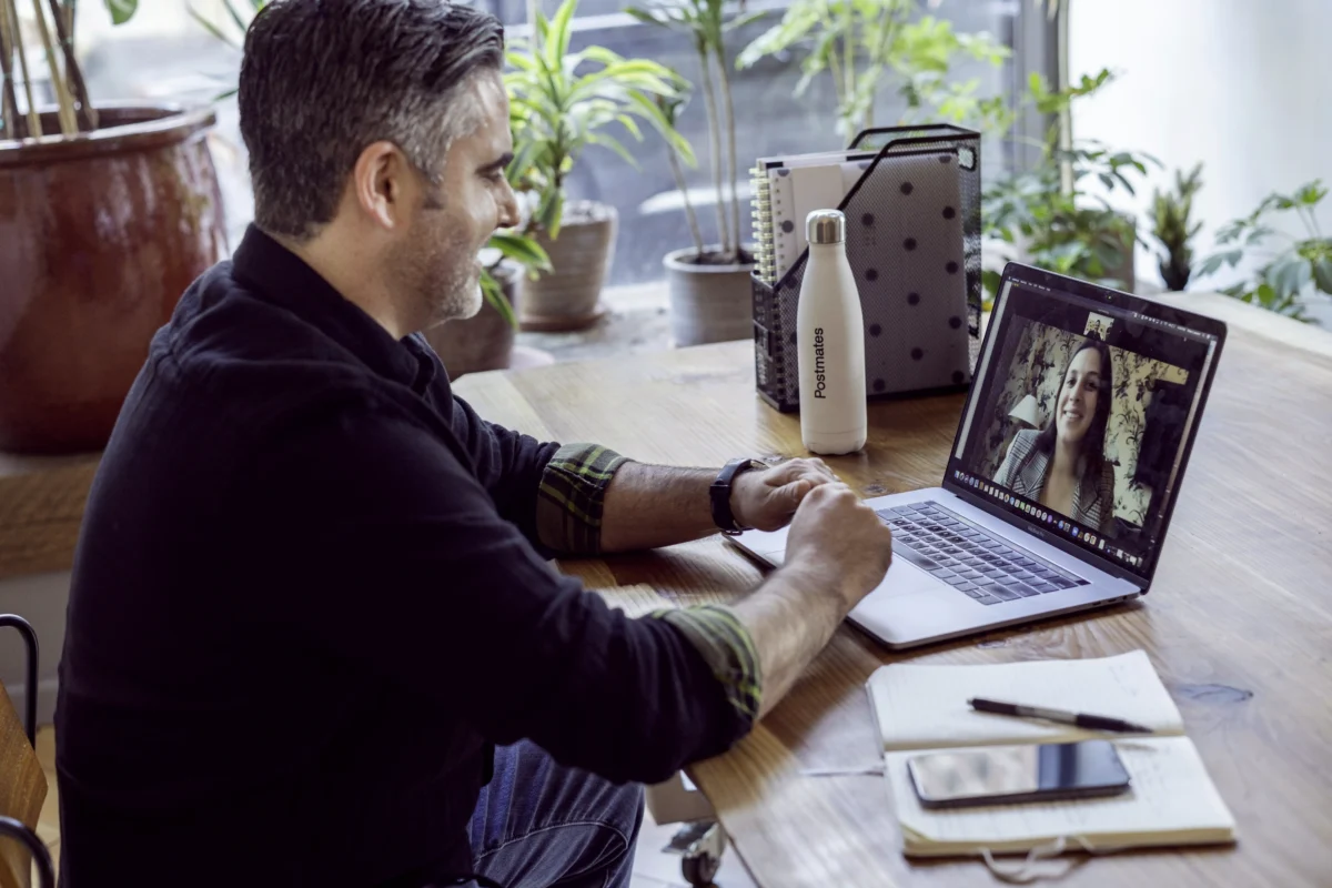 Homme en visioconférence avec une femme via un ordinateur portable, accompagné d'objets de bureau sur une table, avec des plantes en arrière-plan.