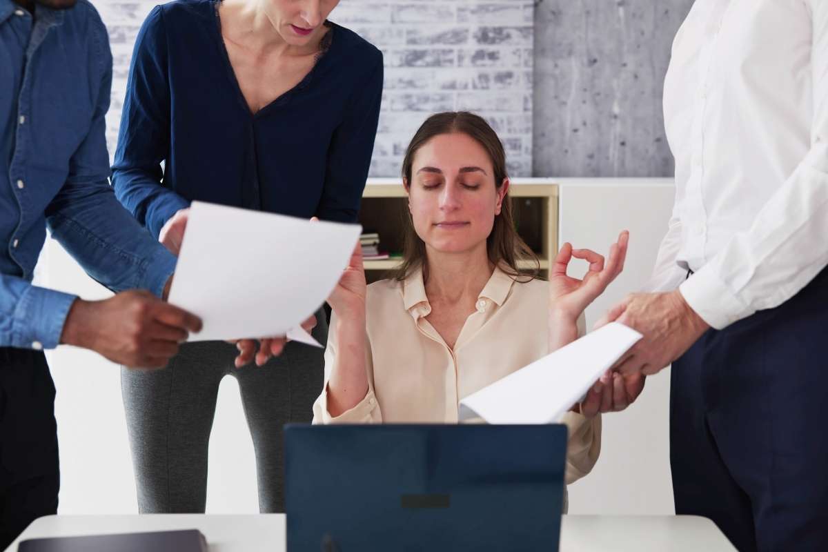 Une femme médite devant un ordinateur portable, entourée de collègues tenant des documents, illustrant la gestion des conflits en équipe.